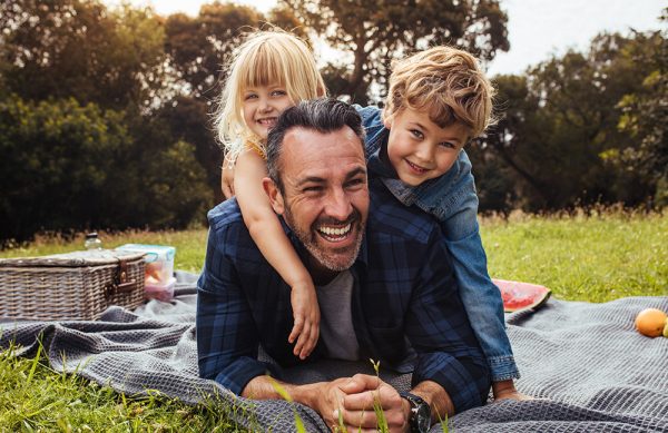 Happy man with kids on a picnic lying down in park beside a picnic basket. Children lying on the back of their father.