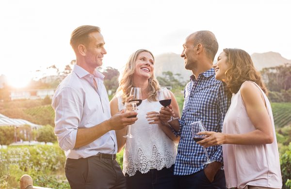 Two relaxed mature couples laughing and enjoy glasses of red wine at sunset. Group of happy friends celebrating outdoor and toasting with glassess. Happy man and smiling women having fun at vineyard.